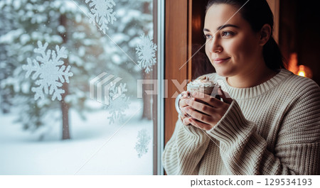 Woman enjoying hot chocolate in winter looking out window with snowflakes. Cozy hygge moment with female holding mug of cocoa on cold snowy day 129534193