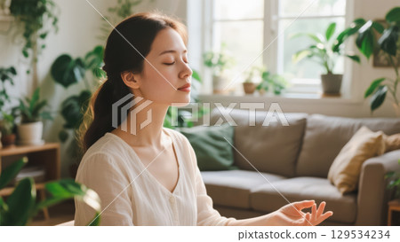 Beautiful young woman enjoying morning meditation in cozy living room, healthy lifestyle concept for mental wellness and peaceful spiritual practice at home 129534234