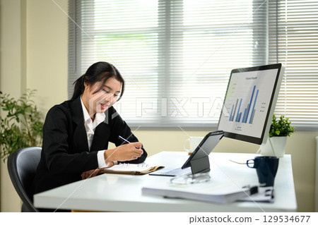 Businesswoman analyzing bar charts and writing in a notebook during a busy work session 129534677