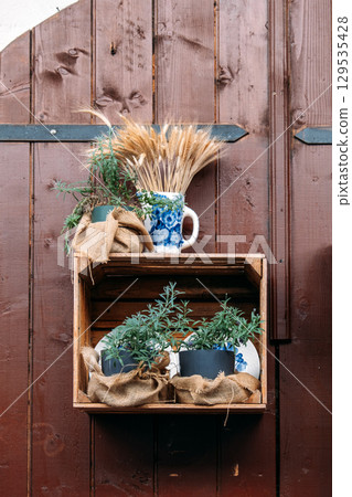 Wooden crate shelf mounted on a rustic wooden door, holding potted herbs wrapped in burlap and a ceramic mug filled with wheat stalks. Cottagecore decor, rustic, vintage kitchen vibes, slow living  129535428