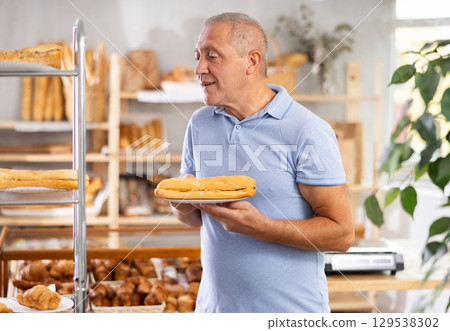 Senior male customer stands with croissant in hands near window of bakery. 129538302