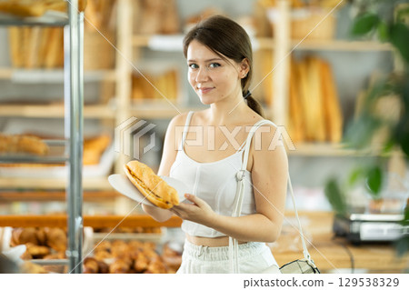 Young female customer stands with bocadillo in hands near window of bakery. Young female customer stands with bocadillo in hands near window of bakery. 129538329