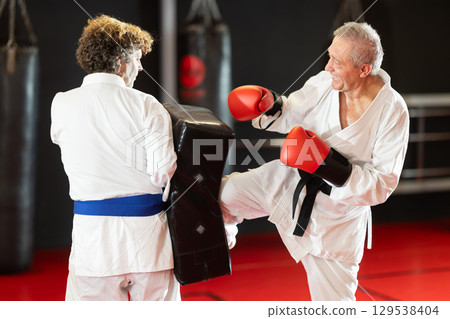 Elderly man in white kimono kicks and punches a punching bag held by trainer Elderly man in white kimono kicks and punches a punching bag held by trainer 129538404