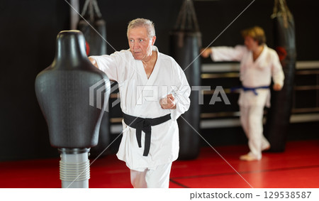 Elderly man in white kimono trains in gym - he punches sports dummy 129538587