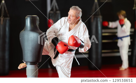 Aged man in white gi practicing strikes on dummy in dojo Aged man in white gi practicing strikes on dummy in dojo 129538651
