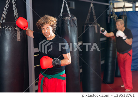 Martial artist in boxing gloves resting against punchbag in dojo Martial artist in boxing gloves resting against punchbag in dojo 129538761