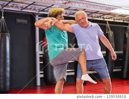 Mature man and adult man practice self-defense techniques at gym 129538918