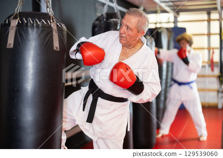 Senior man in white gi training with punching bag in dojo Senior man in white gi training with punching bag in dojo 129539060
