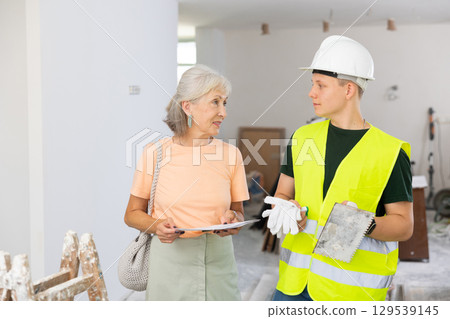 Mature woman discussing room renovation work plan with guy in protective helmet anf yellow vest 129539145