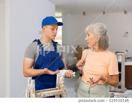 Young builder in blue overalls and an elderly woman are discussing the renovation of room in an apartment Young builder in blue overalls and an elderly woman are discussing the renovation of room in an apartment 129539146