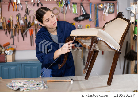 Professional craftswoman restorer in uniform renewing old-fashioned chair while working in repair shop 129539157