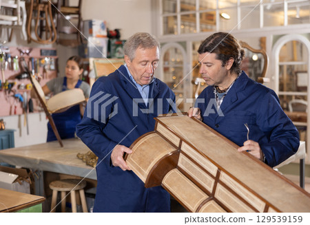 Senior male furniture workshop worker and his apprentice repairing vintage commode 129539159