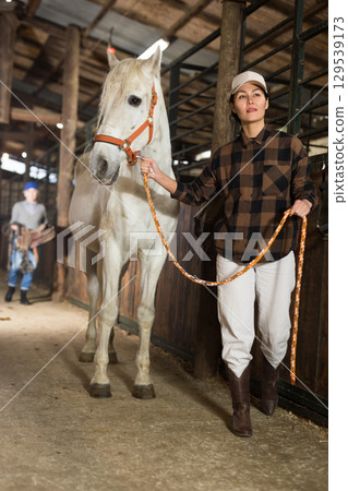 Asian woman leads a white horse by the bridle from stable 129539173