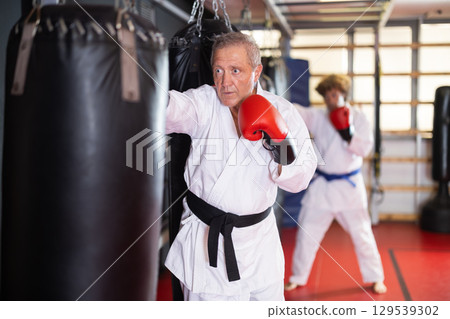 Elderly man in white kimono practices punching punching bag Elderly man in white kimono practices punching punching bag 129539302