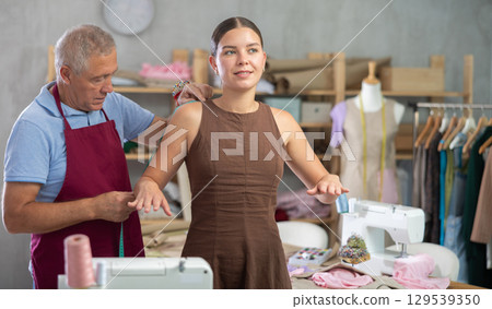 Elderly man tailor takes body measurements from young woman 129539350
