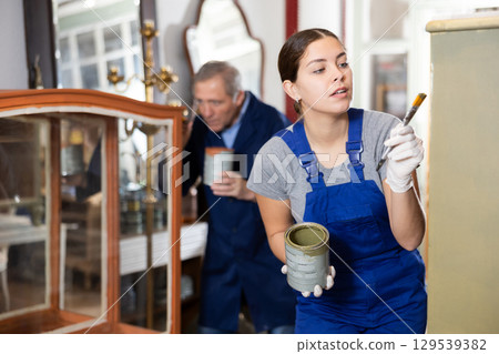 Skilled female furniture repair specialist in uniform painting furniture during workday in workshop 129539382