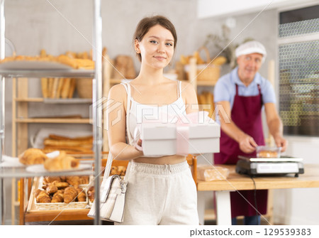 Cute young girl customer holding box with decorative bow and buying sweet authors flour products in bakery shop 129539383