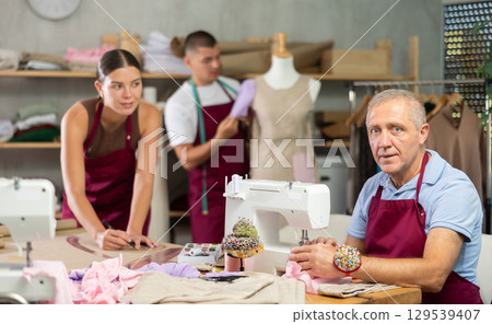 elderly worker sews clothes against the background of young workers in the atelier elderly worker sews clothes against the background of young workers in the atelier 129539407