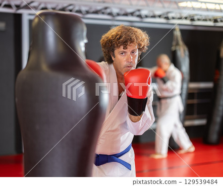 Man in white kimono trains in gym - he punches sports dummy Man in white kimono trains in gym - he punches sports dummy 129539484