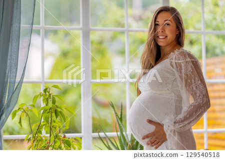 Pregnant woman in a white dress smiling near bright window and plants Pregnant woman in a white dress smiling near bright window and plants 129540518