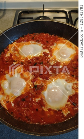 Shakshuka with eggs poached in a rich tomato sauce with onions and herbs cooks in a frying pan 129543793