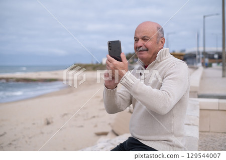 An elderly person sitting by the ocean shore, taking photos of the view.  129544007