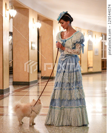 Elegant woman in blue dress walking white fluffy Pomeranian dog indoors Elegant woman in blue dress walking white fluffy Pomeranian dog indoors 129544074