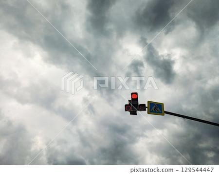 Red traffic light and pedestrian crossing sign against a gloomy cloudy sky. 129544447