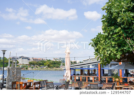 an outdoor seaside cafe with wooden tables and chairs, shaded by large umbrellas and a tree 129544707