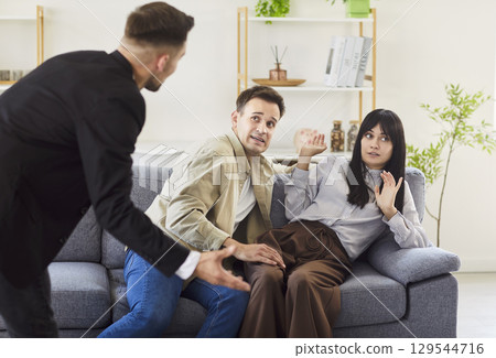Angry man confronts anxious couple on sofa during tense home argument scene Angry man confronts anxious couple on sofa during tense home argument scene 129544716