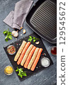 Raw meat sausages on a slate board, with herbs and spices and a square grill pan. View from above 129545672