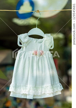 Tender kids clothes on hanger, summer warm rays. Elegant summer white baby dress with lace trim and decorative fabric roses hanging in display. Concept of childhood, innocence, delicate fashion 129545681