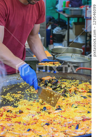 Street Paella Cooking. Chef in red shirt serving seafood paella into takeaway box at outdoor street food market with large pan. Concept of paella cooking, street food, authentic Spanish cuisine 129546099