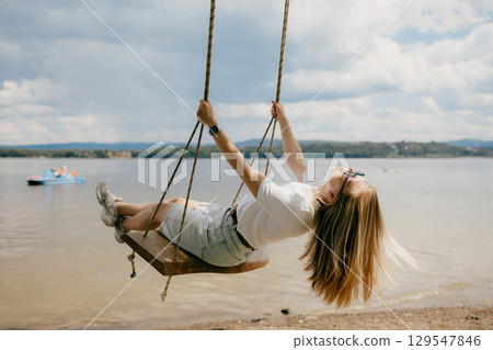 Happy young woman wearing sunglasses and casual clothes swings joyfully over serene lake, embracing warmth of summer holidays and feeling of freedom. Woman in casual attire swinging near tranquil lake Happy young woman wearing sunglasses and casual clothes swings joyfully over serene lake, embracing warmth of summer holidays and feeling of freedom. Woman in casual attire swinging near tranquil lake 129547846