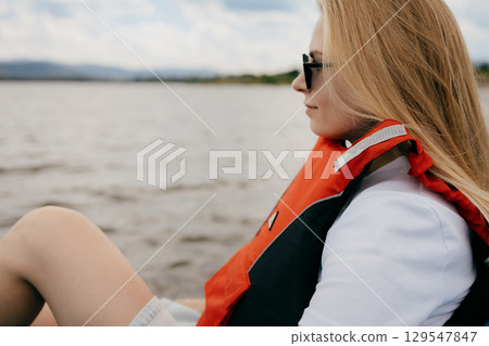 Young blonde woman wearing sunglasses and life jacket, enjoying scenic boat trip on tranquil lake while contemplating beautiful landscape and serene surroundings. Blonde woman relaxing in life jacket 129547847