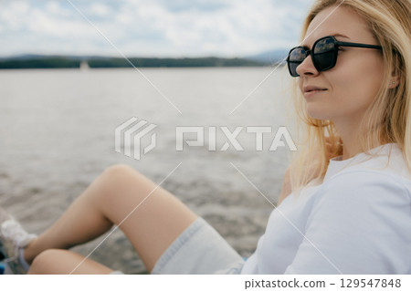 Young blonde woman wearing stylish sunglasses, relaxing by serene lake shore and enjoying blissful summer vacation, with sailboat gracefully sailing in background. Millennial woman on water bike  129547848