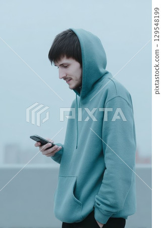 Young male with cellphone against blue wall. Fresh vibe for chatting or social media themes, ideal for digital or modern designs. Great for youth or tech visuals. 129548199