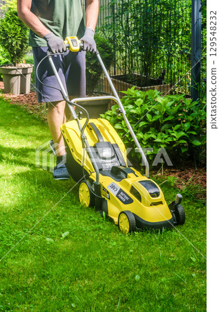Close-up of a person pushing a bright yellow battery-powered lawn mower while cutting grass in a sunny garden. High quality photo 129548232