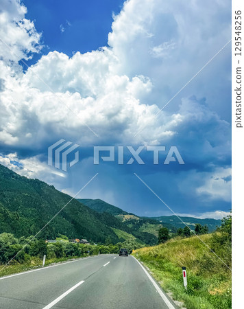 Beautiful two-lane mountain road stretching into the distance with lush green hills and dramatic white clouds. High quality photo 129548256