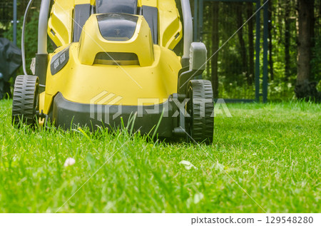 Low angle close-up of a bright yellow electric lawn mower trimming grass in a green garden. High quality photo Low angle close-up of a bright yellow electric lawn mower trimming grass in a green garden. High quality photo 129548280