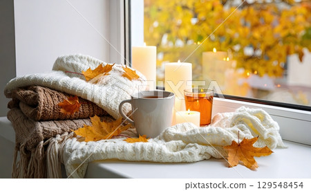 A window with a white blanket and a cup of tea on top of it. The blanket has leaves on it and the cup is filled with tea. There are candles on the table as well 129548454