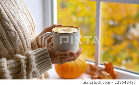 A woman holding a white coffee mug with a pumpkin on the table. The pumpkin is orange and the woman is wearing a white sweater A woman holding a white coffee mug with a pumpkin on the table. The pumpkin is orange and the woman is wearing a white sweater 129548455