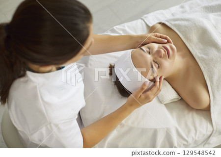Beauty care session in clinic, woman lying on treatment bed with eyes closed, beautician hands 129548542