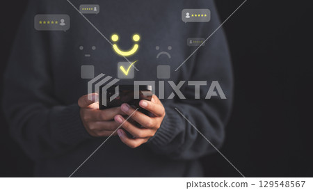 Female customer using a smartphone select a happy face icon on digital feedback. Concept of customer satisfaction, testimonial, online reviews, user feedback, review rankings and service quality. Female customer using a smartphone select a happy face icon on digital feedback. Concept of customer satisfaction, testimonial, online reviews, user feedback, review rankings and service quality. 129548567