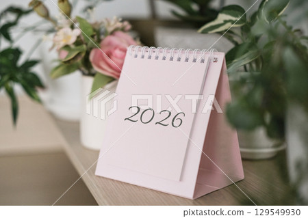 Desk calendar for year 2026 on wooden shelf surrounded by lush green plants and soft-pink flowers, suggesting planning and fresh start 129549930