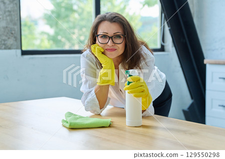 Smiling beautiful middle aged woman in gloves with rag cleaning spray looking at camera Smiling beautiful middle aged woman in gloves with rag cleaning spray looking at camera 129550298