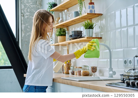 Young woman in rubber gloves cleaning kitchen, wiping dust with rag 129550305