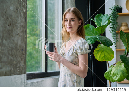 Young beautiful smiling romantic girl looking at camera with cup, standing near window Young beautiful smiling romantic girl looking at camera with cup, standing near window 129550329
