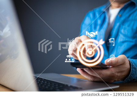 A businessman in a blue shirt interact with a smartphone, displaying holographic business icons, including a glowing target symbolizing goal achievement, analytics, partnerships, and financial growth. 129551329
