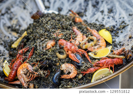 Close-up of black rice seafood paella with crayfish, shrimp, squid, mussels, and lemon wedges in large pan. Concept of black paella, seafood delicacy, Mediterranean gourmet cuisine 129551812